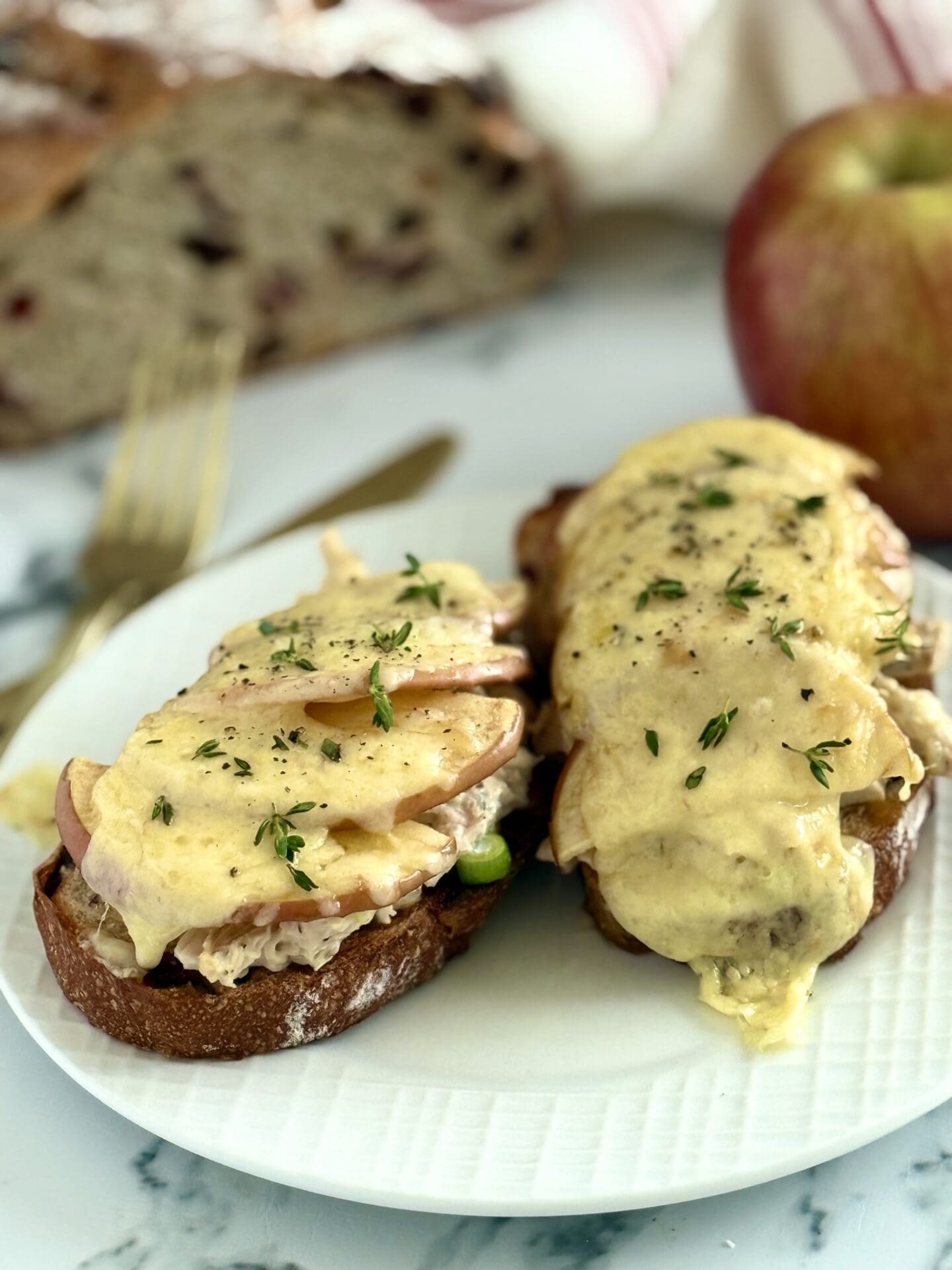 IMG_6906 Chicken and Apple Melt open-faced sandwiches are seen on a white plate. The smoked cheddar cheese is melted over the tops and drips down the sides of the sandwiches that are stacked high with chicken salad and apple slices.