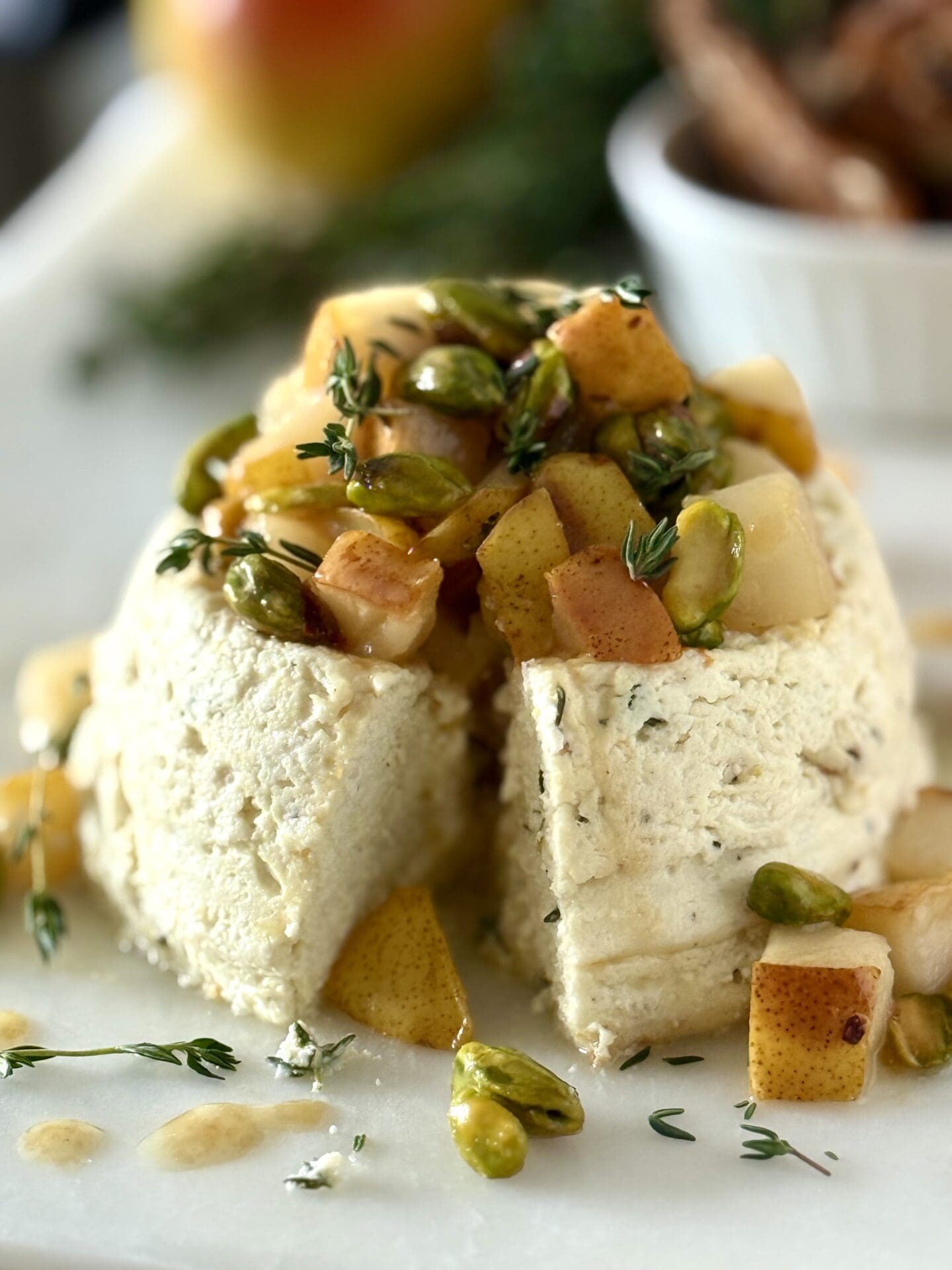 A Pistachio and Pear Baked Ricotta appetizer sits on a white marble serving platter. A wedge of the baked cheese has been cut away to revel the creamy centre.