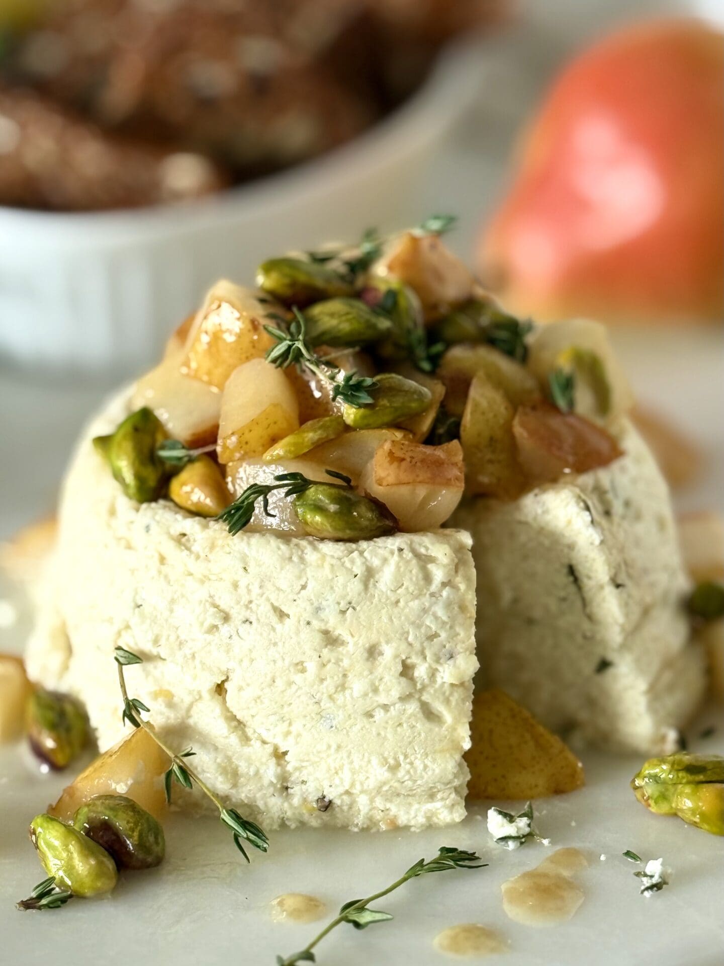 A mini cheesecake made of ricotta, lemon and thyme sits on a marble platter, covered with caramelized pistachio and pear topping. A slice of the cheesecake has been removed revealing the creamy centre. Crackers and fresh pears are seen in the background.