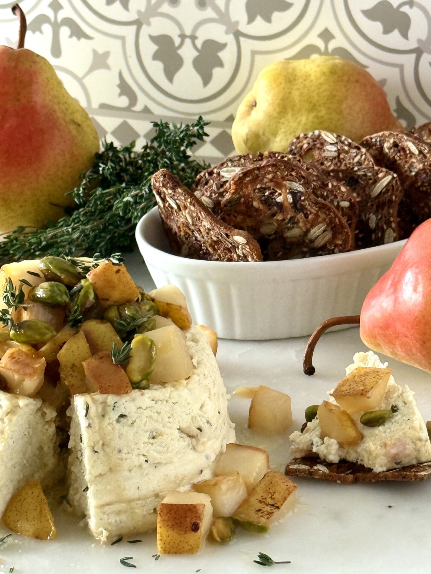 Pistachio and Pear Baked Ricotta is shown, lavishly garnished with caramelized pears, green pistachios and sprigs of fresh time. The appetizer is surrounded by crisp crackers for serving and red and yellow pears.