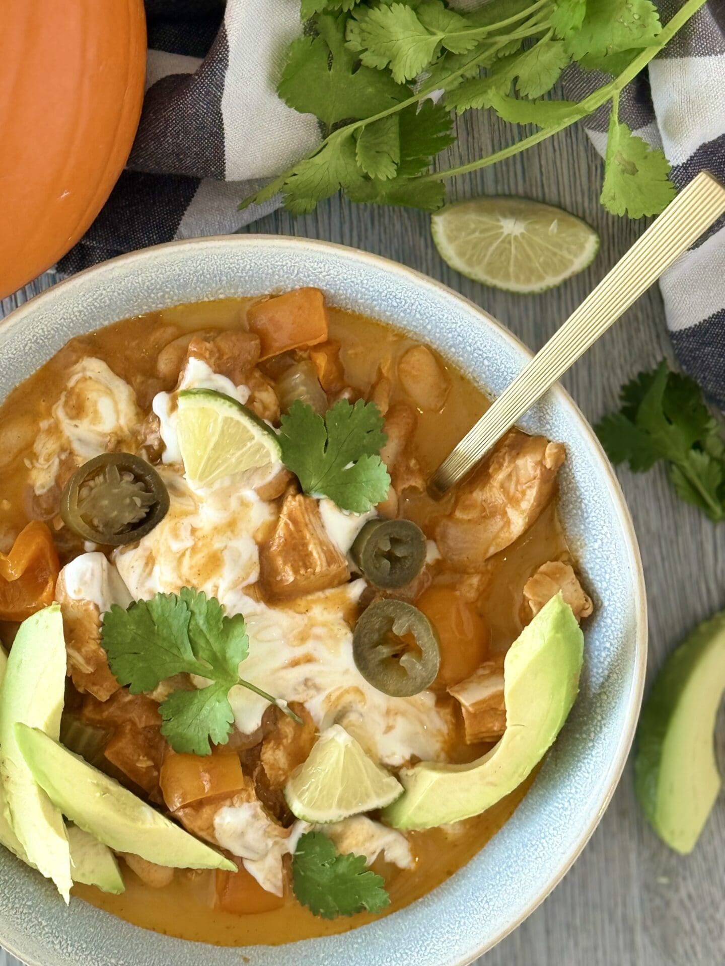 A bowl of Pumpkin Chicken Chili is seen from above, generously garnished with avocado slices, jalapeño, lime, cilantro and sour cream.