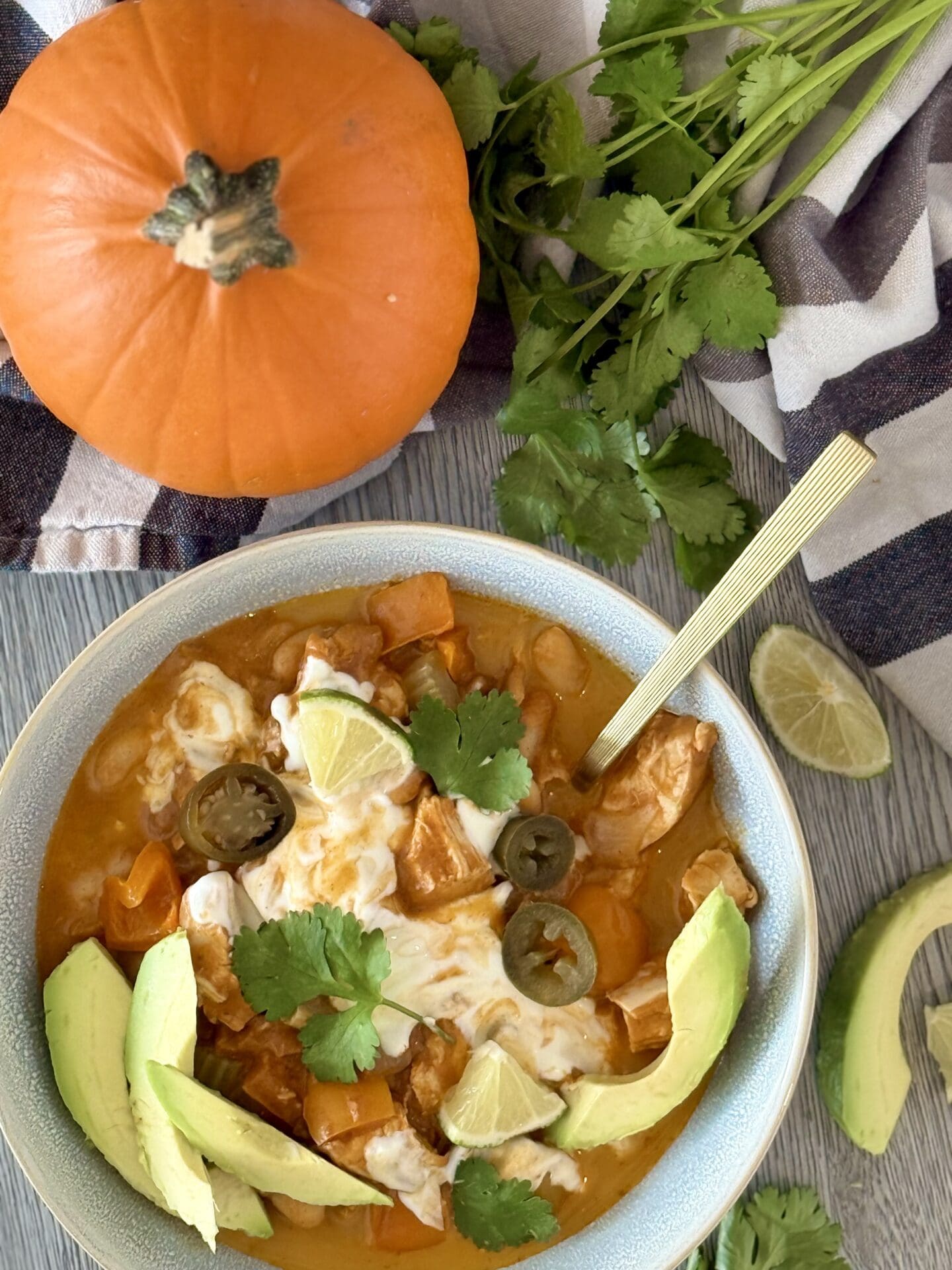 A bowl of Pumpkin Chicken Chili is seen from above, surrounded by fresh cilantro, avocado slices, lime wedges and a small orange pumpkin.