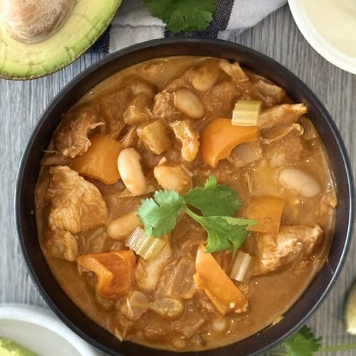 A bowl of Pumpkin Chicken Chili is seen from above, garnished with fresh cilantro. The bowl is surrounded by garnishes such as fresh avocado, sour cream and lime wedges.