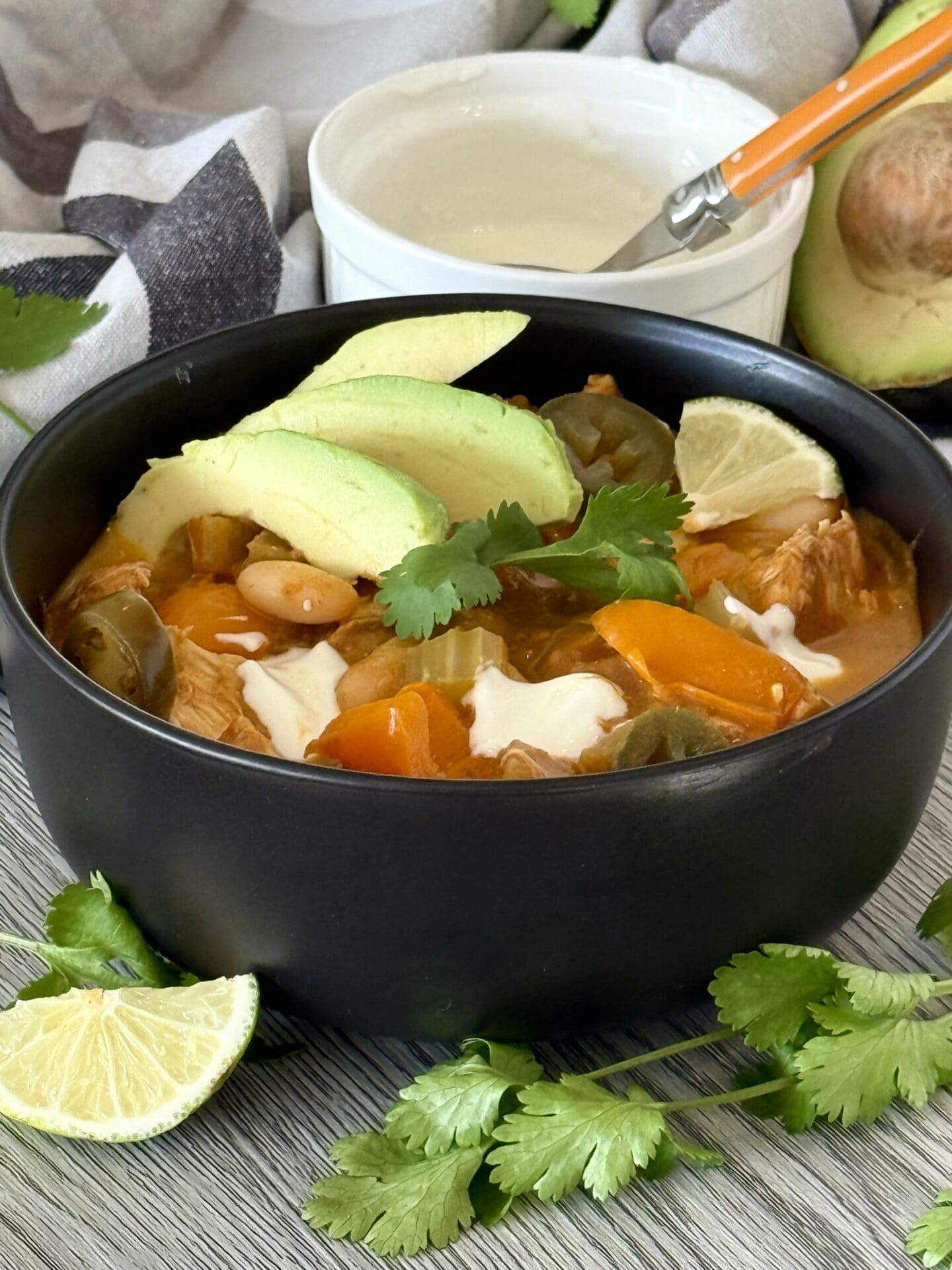 A black bowl filled with garnished Pumpkin Chicken Chili sits on a grey wood table. A bowl of sour cream and a avocado half are seen in the background.