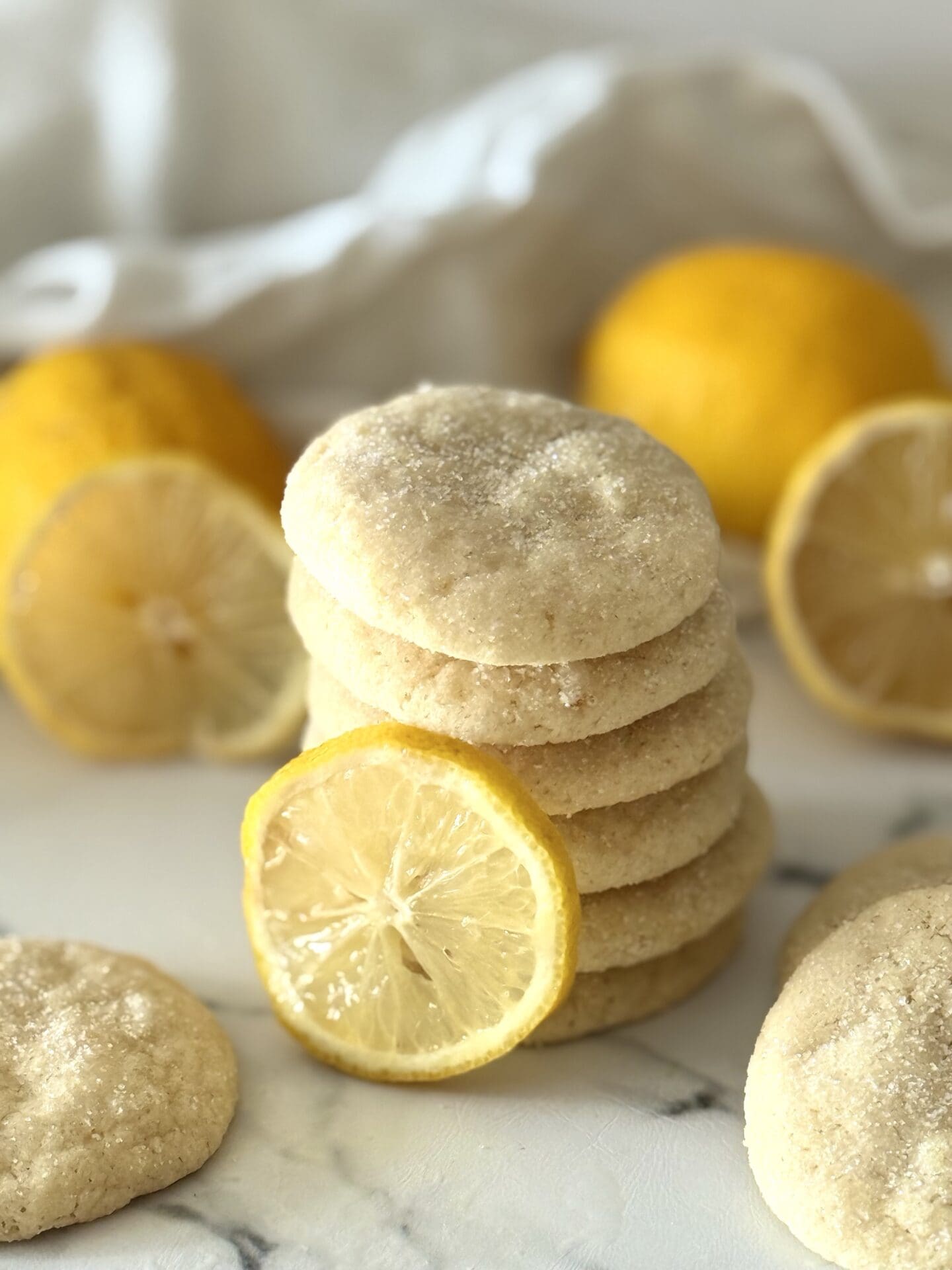 A stack of glittering chewy lemon sugar cookies is seen on a white marble countertop, surrounded by whole lemons and fresh lemon slices.