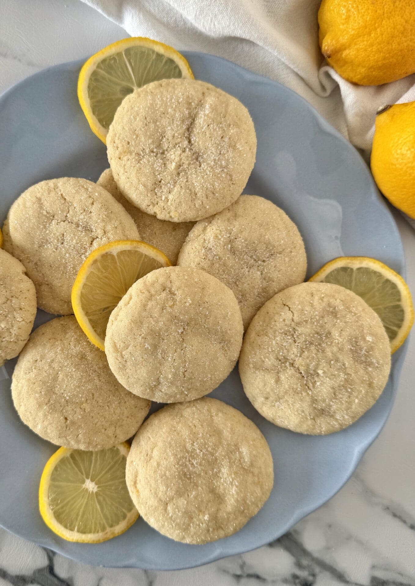 A light blue plate is filled with soft and chewy lemon sugar cookies.  Slices of fresh lemon are tucked around the cookies.