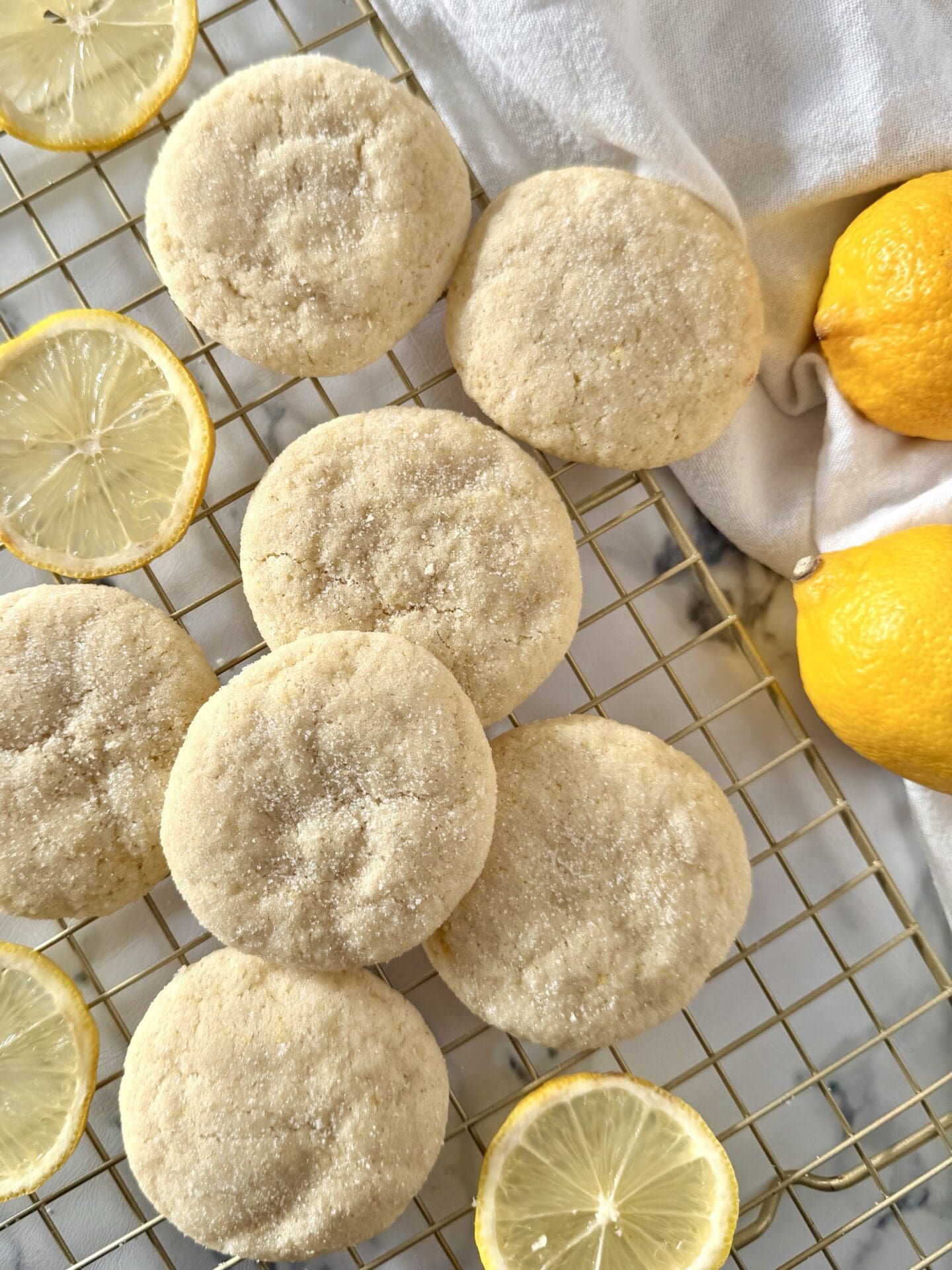 Freshly baked soft and chewy lemon sugar cookies are seen from above, cooking on a gold wire rack, surrounded by lemon slices.