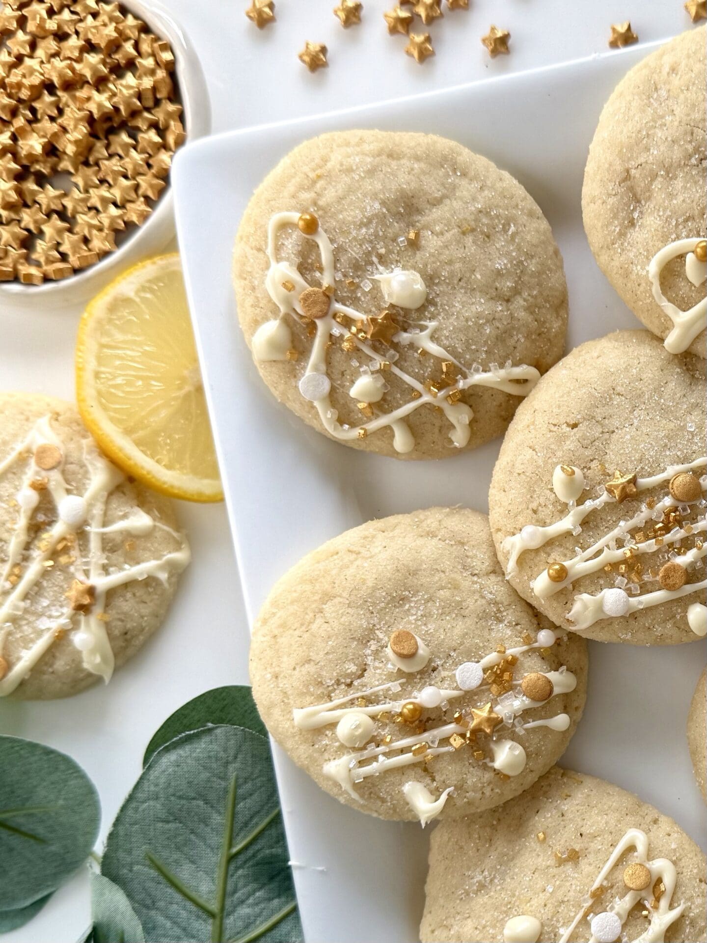 A plate of soft and chewy lemon sugar cookies decorated with white chocolate and sprinkles sits on a platter.  Lemon slices, greenery and a dish of star candies completes the festive scene.