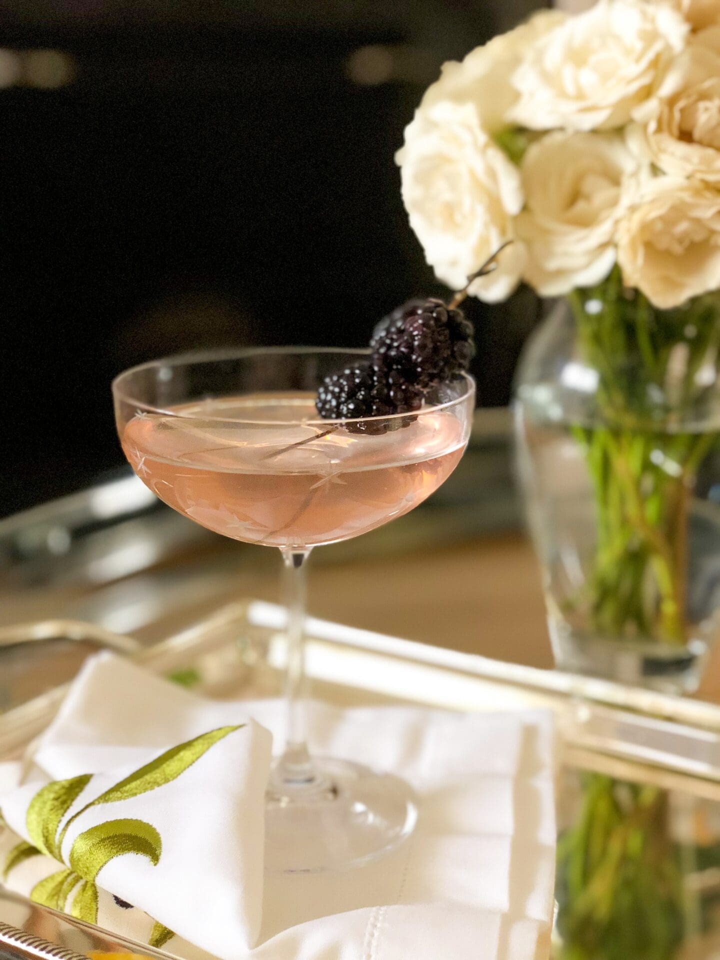 A pretty pink Blackberry Blush Cocktail is served in an elegant coupe glass on a silver tray. A vase of white roses appears in the background.