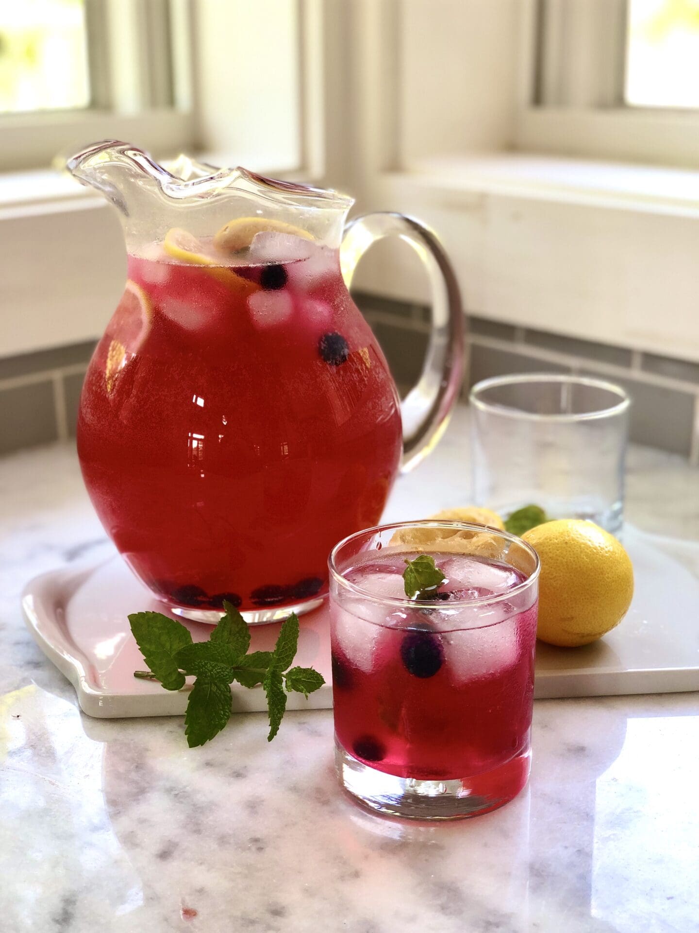 A pitcher of ice cold blueberry lavender lemonade sits on a white marble counter by a window.  A glass of the drink sits in the foreground, garnished with lemon slices and fresh blueberries.