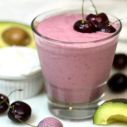 A glass of pink cherry vanilla smoothie is seen on a white marble counter. The glass is garnished with fresh cherries and is surrounded by fresh avocado and more cherries.
