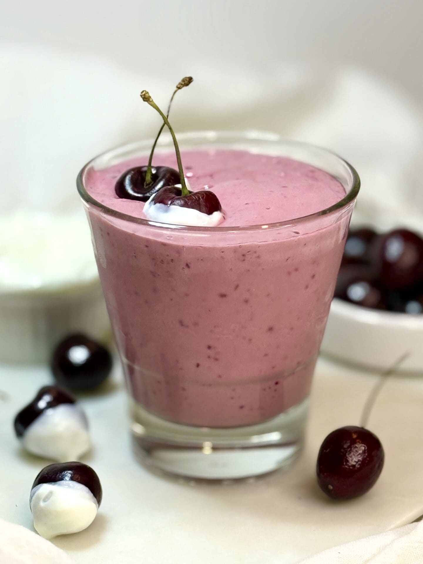 A glass of pink cherry vanilla smoothie is seen on a white marble counter surrounded by yogurt dipped cherries.