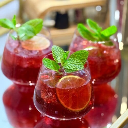 Three glasses of Pomegranate Lime Mocktail are garnished with fresh mint, lime wheels and pomegranate arils. The glasses sit together on a mirrored bar cart, which shows their reflection.