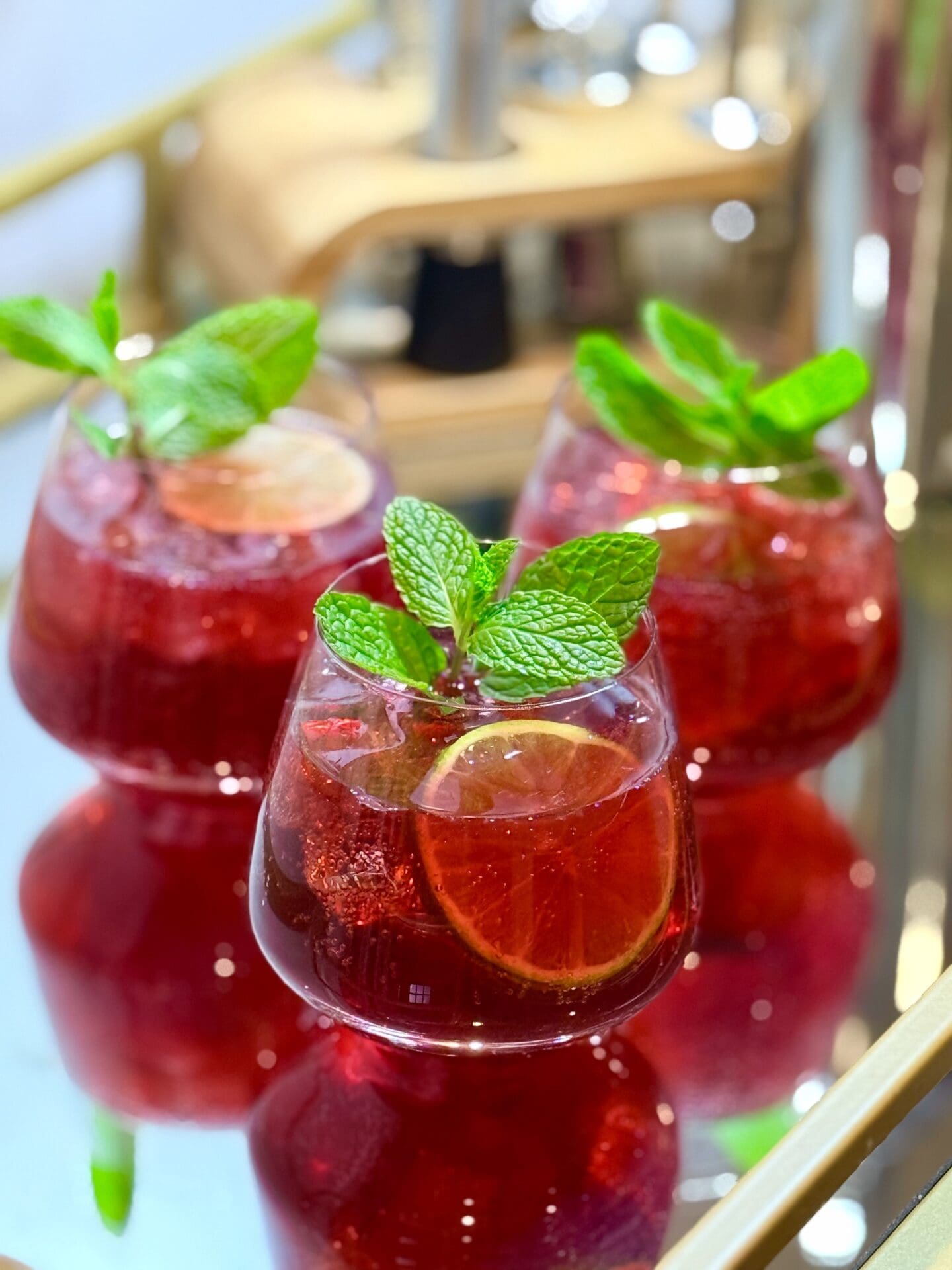 Three glasses of Pomegranate Lime Mocktail are garnished with fresh mint, lime wheels and pomegranate arils. The glasses sit together on a mirrored bar cart, which shows their reflection.