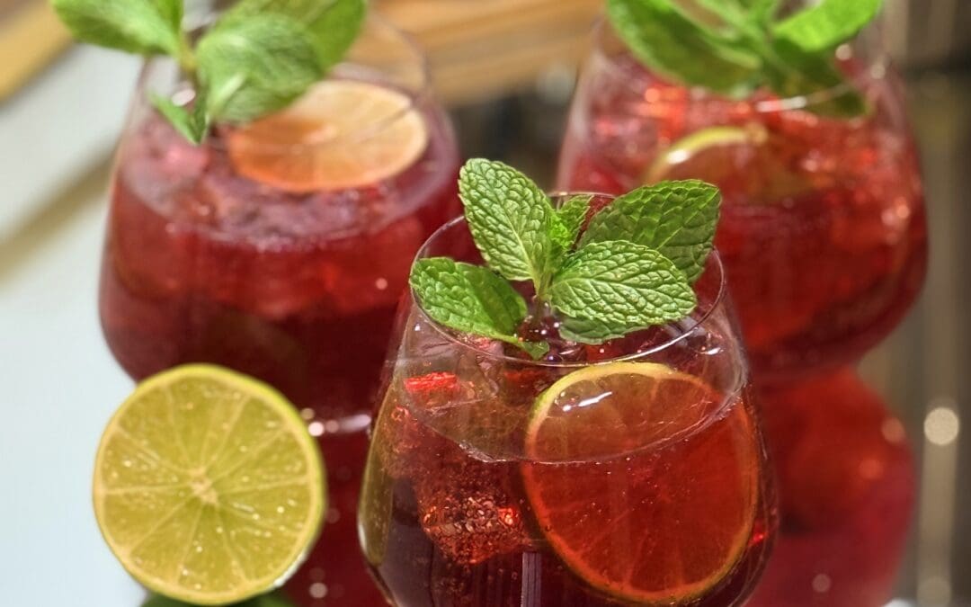 Three beautifully garnished glasses of Sparkling Pomegranate Lime Mocktail sit on a glass and gold bar cart. Bart tender tools can be seen in the background.