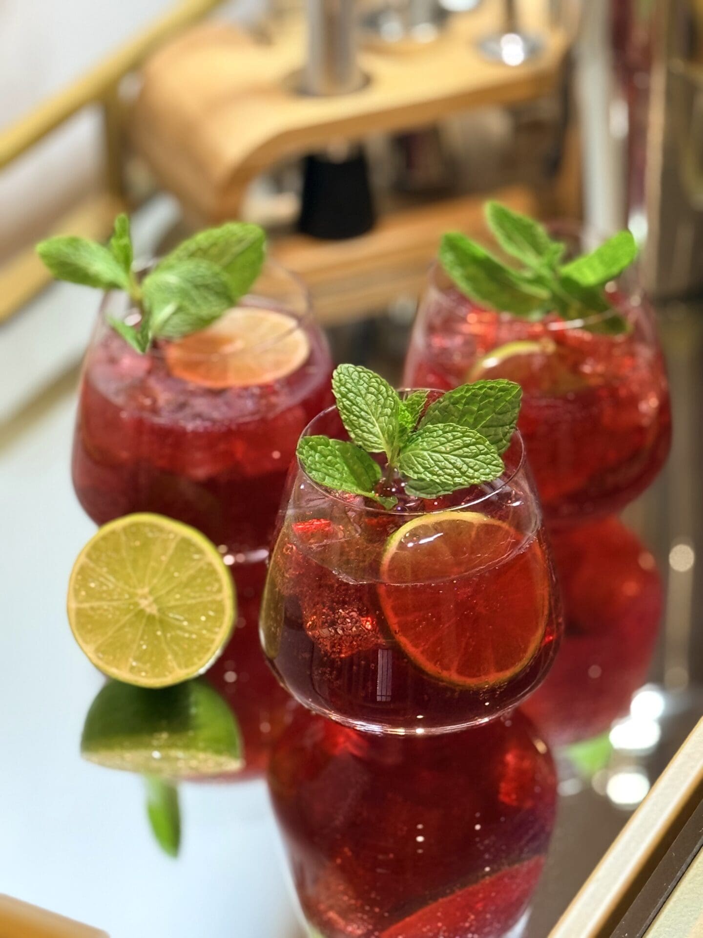 Three beautifully garnished glasses of Sparkling Pomegranate Lime Mocktail sit on a glass and gold bar cart. Bart tender tools can be seen in the background.