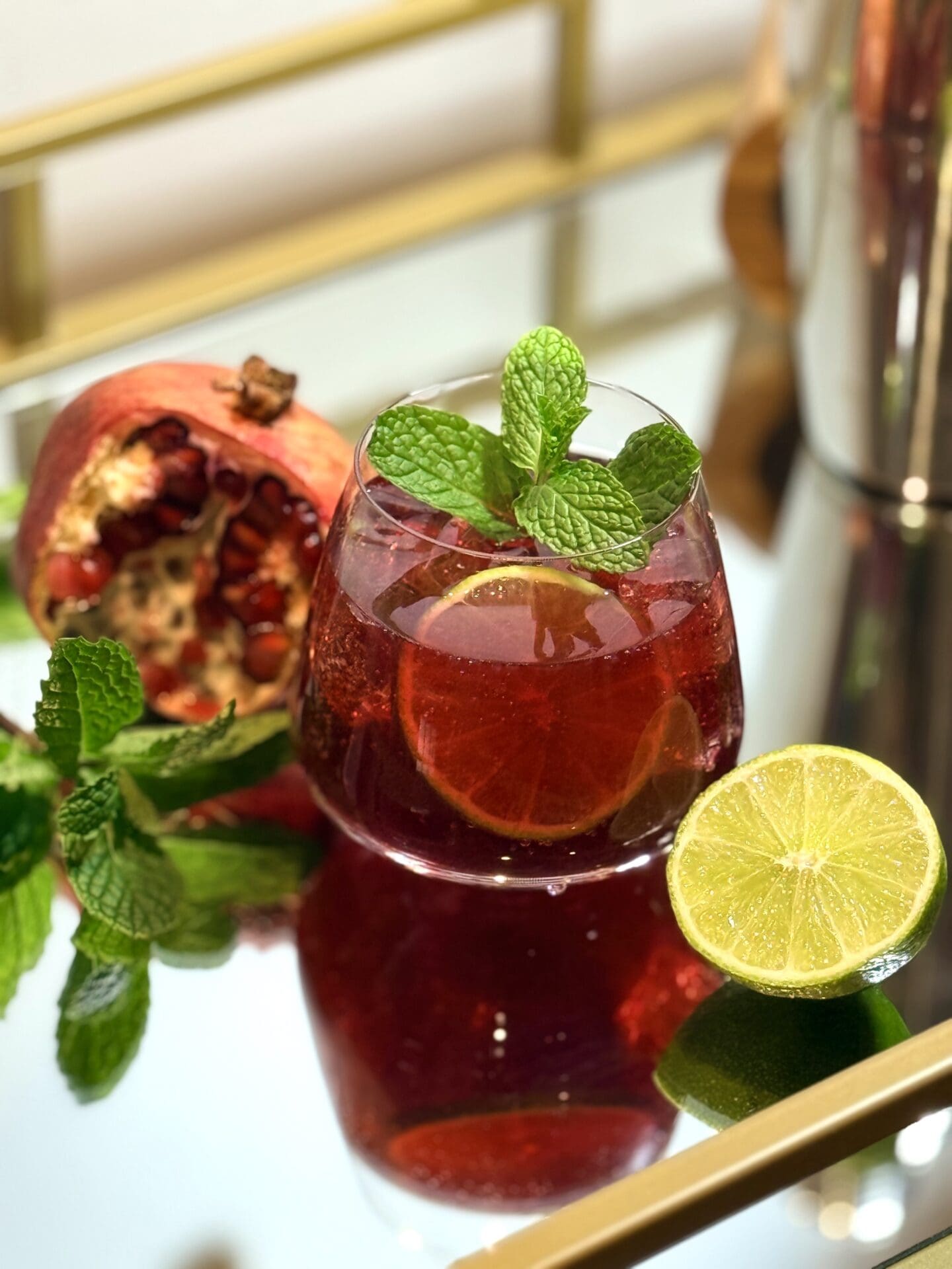 A glass of alcohol-free Sparkling Pomegranate Lime drink sits on a mirrored bar cart, surrounded by a fresh pomegranate, cut lime and sprigs of fresh mint.
