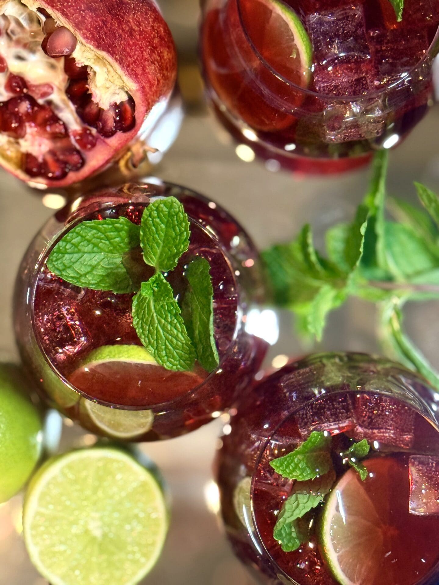 Glasses of ruby red pomegranate lime mocktail are seen from above, garnished with fresh mint, lime and pomegranate.