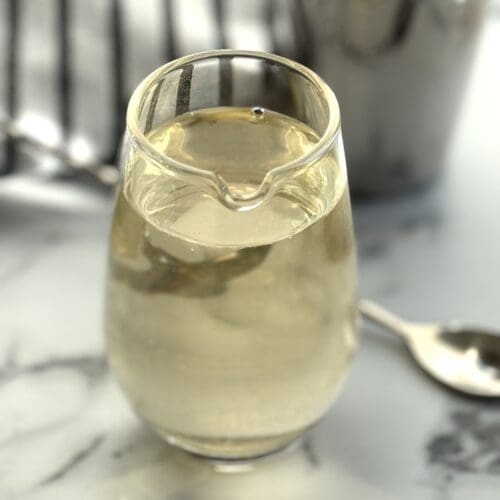 An elegant glass beaker of simple syrup sits on a white marble counter top. A stirring spoon and silver cocktail shaker are seen in the background.