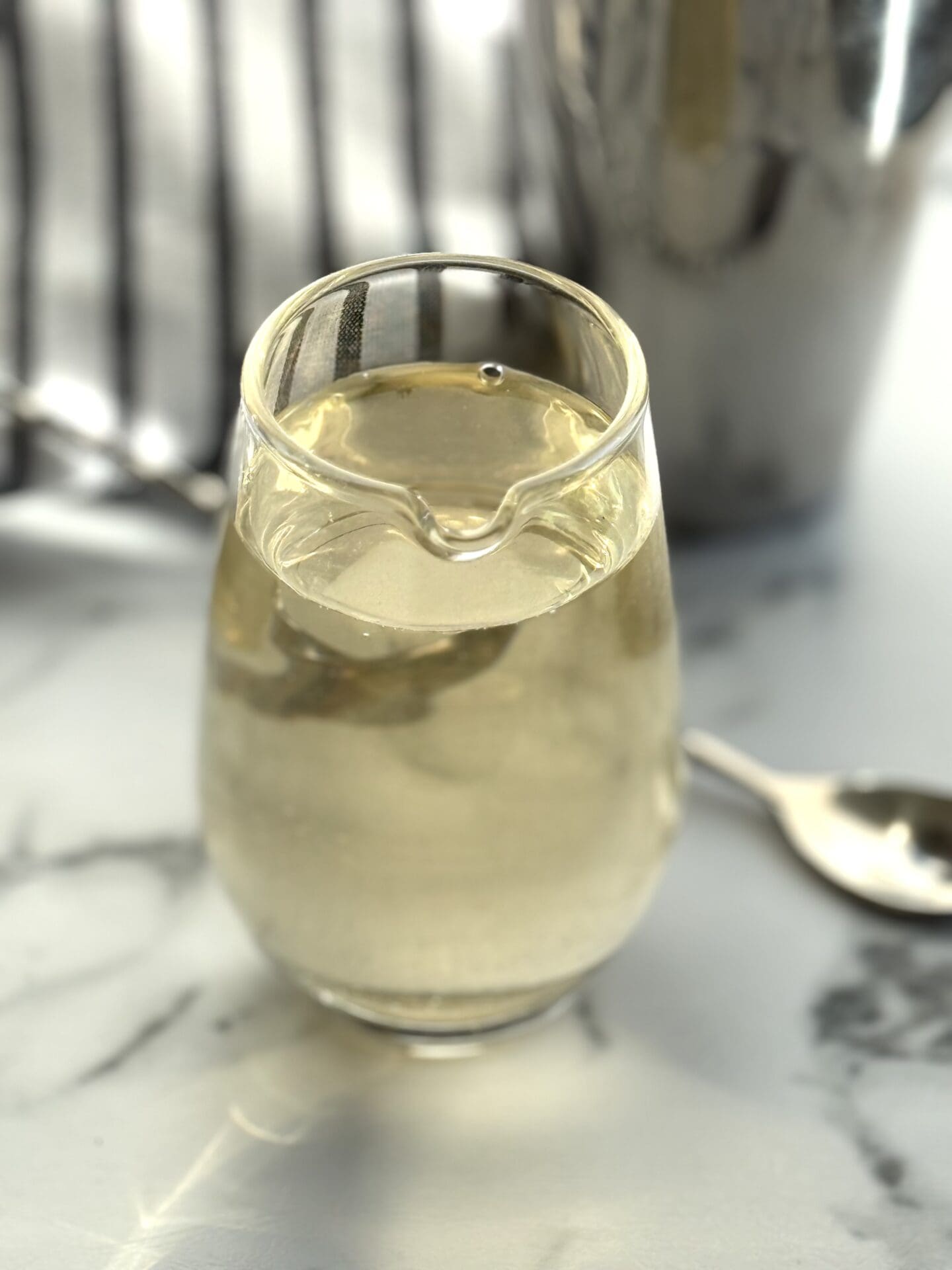 An elegant glass beaker of simple syrup sits on a white marble counter top. A stirring spoon and silver cocktail shaker are seen in the background.