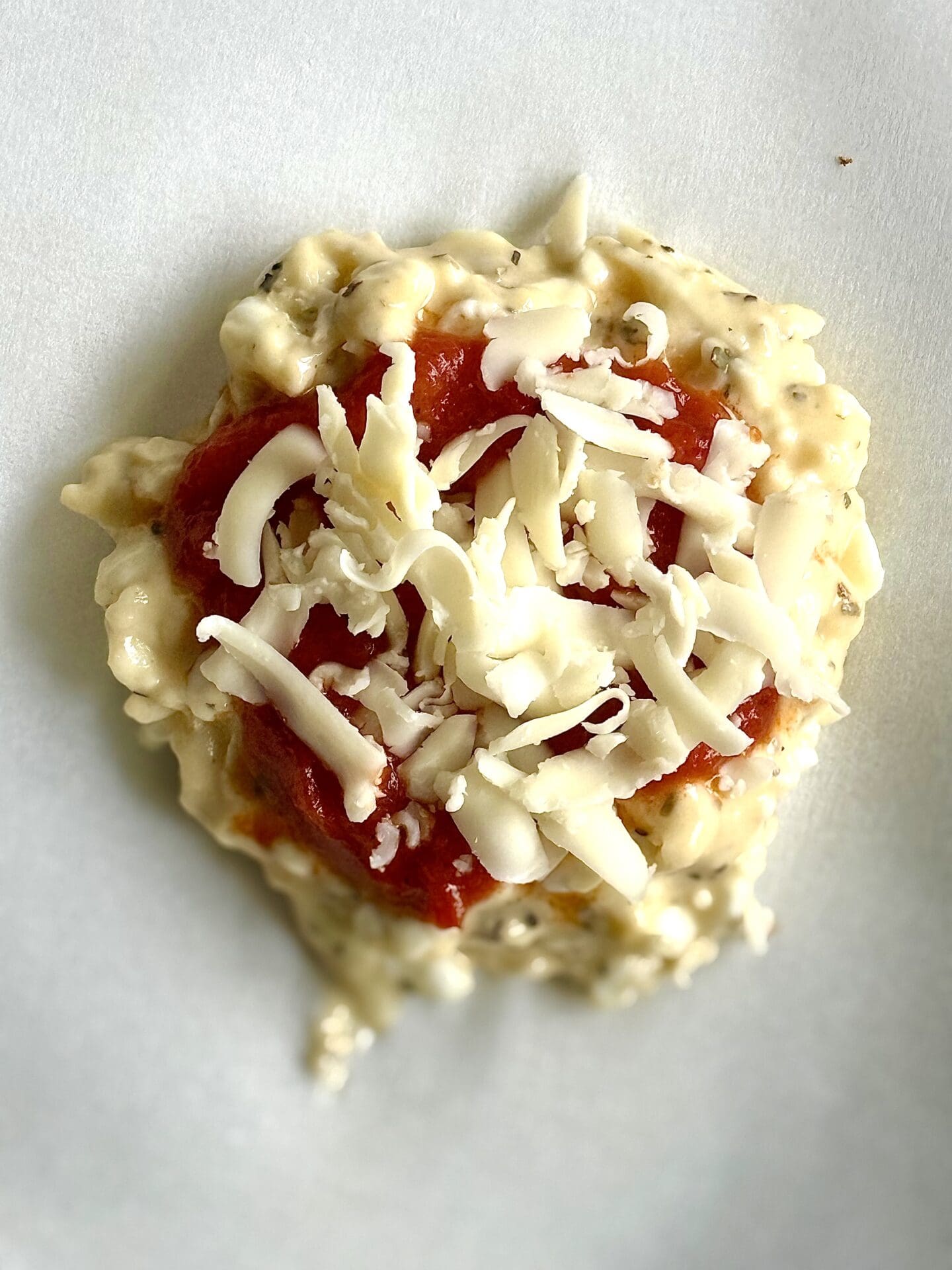 A single unbaked cottage cheese pizza bun is seen from above, showing the assembly of raw batter, marinara sauce and shredded mozzarella cheese.