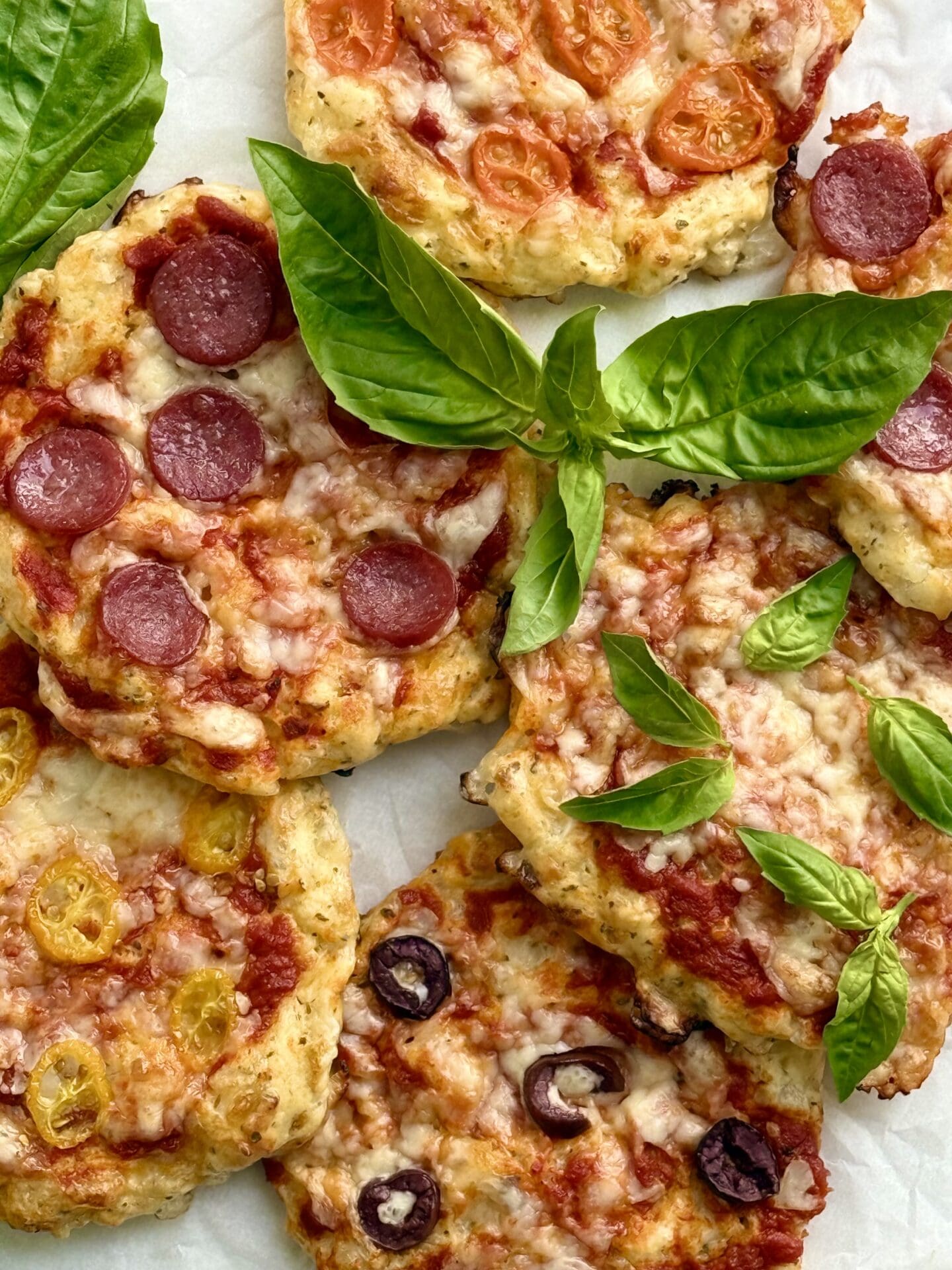 Freshly baked pizza buns with assorted toppings are crowded together on a platter and garnished with fresh basil.