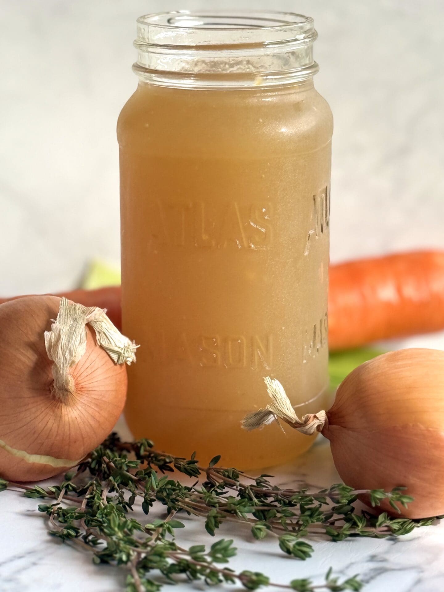 A glass jar of homemade chicken stock sits on a marble counter, surrounded by carrots, celery, onions and  fresh thyme.