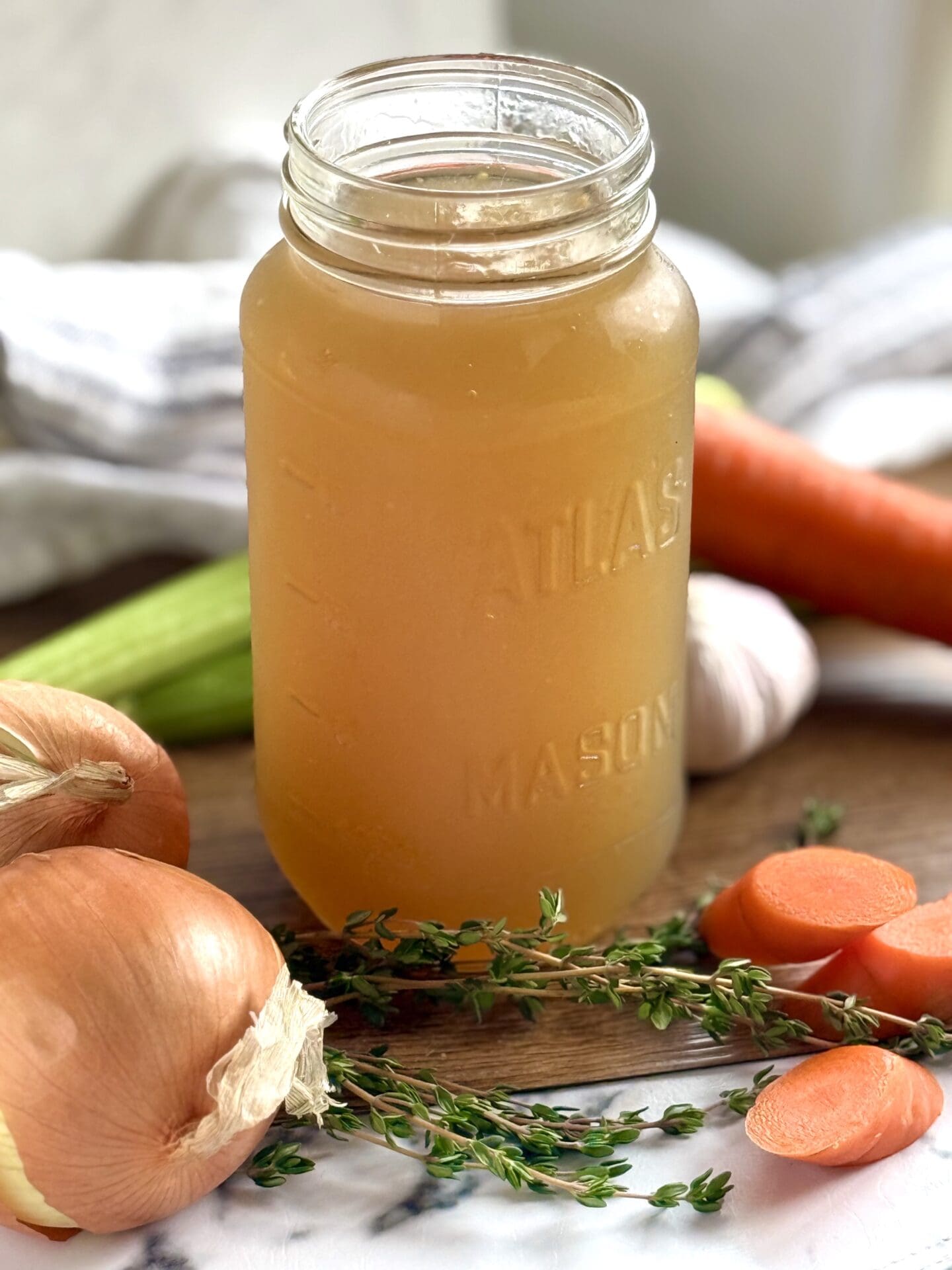 IMG_8164 A jar of golden homemade chicken stock sits on a table surrounded by fresh vegetables and herbs