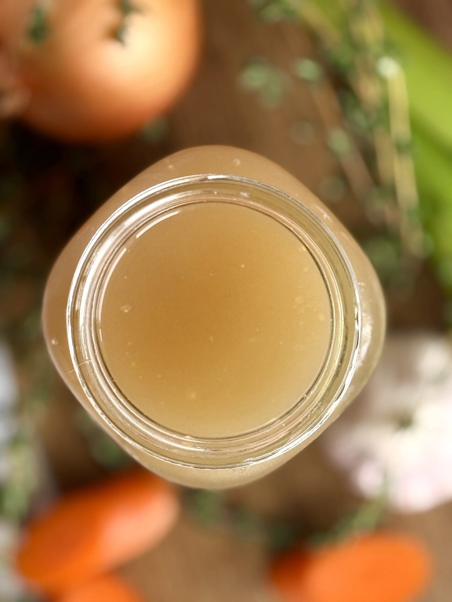 A jar of homemade chicken stock is seen from above, sitting on a wood table surrounded by fresh vegetables and herbs.