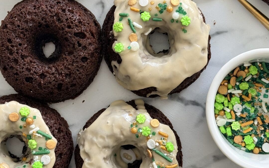 A group of baked chocolate donuts is seen from above. Some are topped with a Baileys glaze and St Patrick’s Day themed sprinkles.