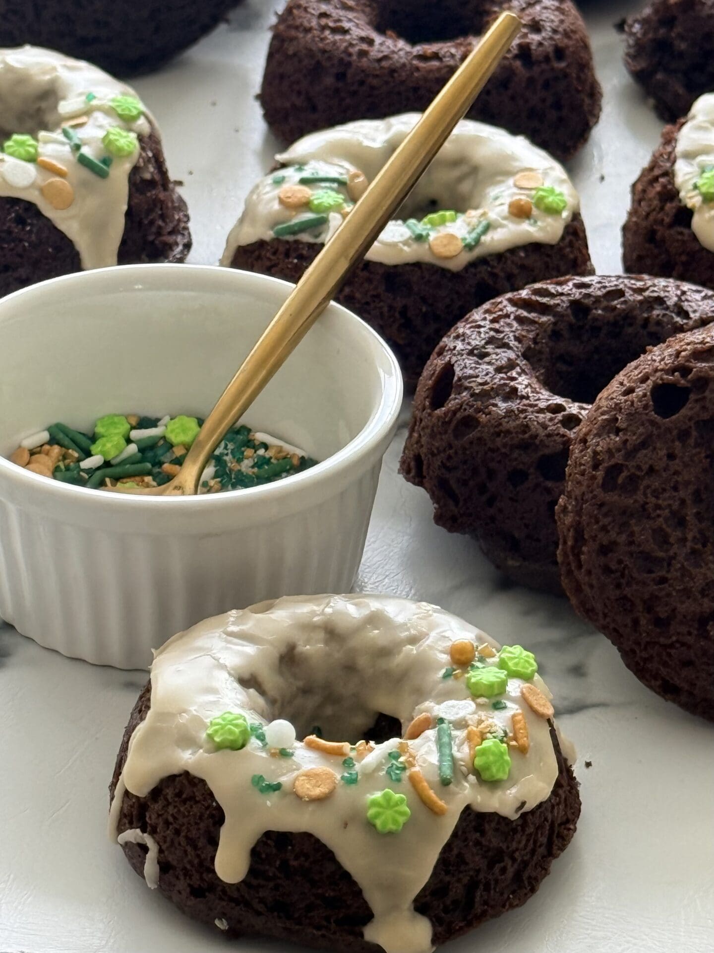 Baked chocolate donuts sit on a countertop, while being topped with a Baileys glaze and green, gold and white sprinkles.