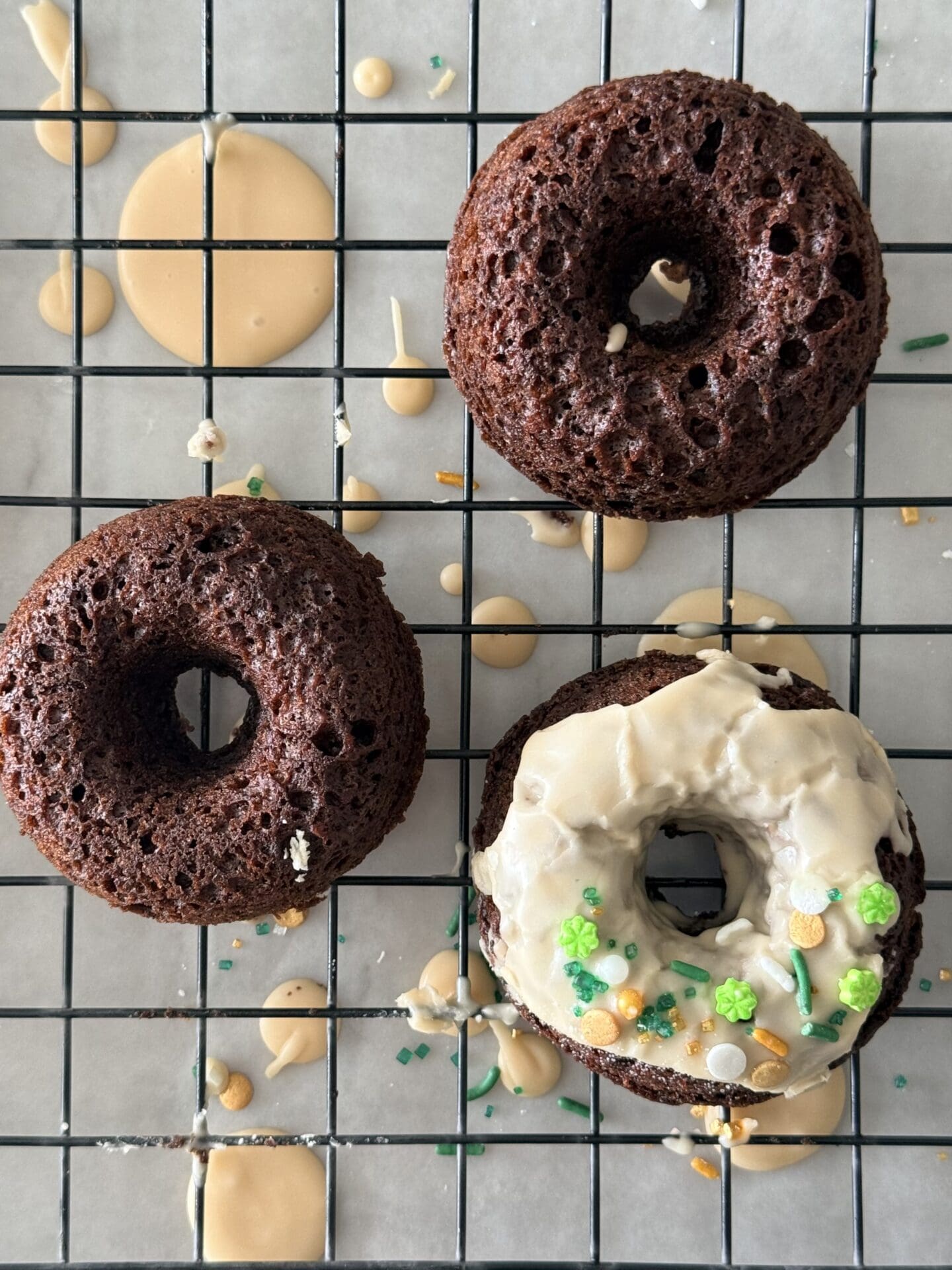 Three baked chocolate donuts sit on a black wire rack. One of the donuts has been topped with a Baileys glaze and St Patrick’s Day sprinkles. Drops of glaze and extra sprinkles can be seen under the rack.