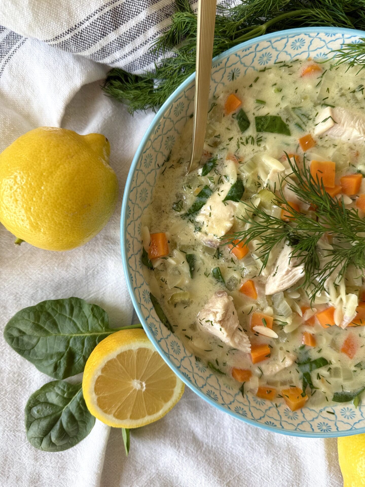 A bowl of Avgolemono soup is seen entering the frame from the right so half of the bowl can be seen in the image.  The soup is surrounded by lemons and baby spinach leaves.