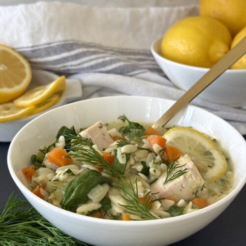 A bowl of Avgolemono soup is seen in the foreground, garnished with fresh dill. Bowls of lemons are seen in the background, along with a blue and white cloth napkin.