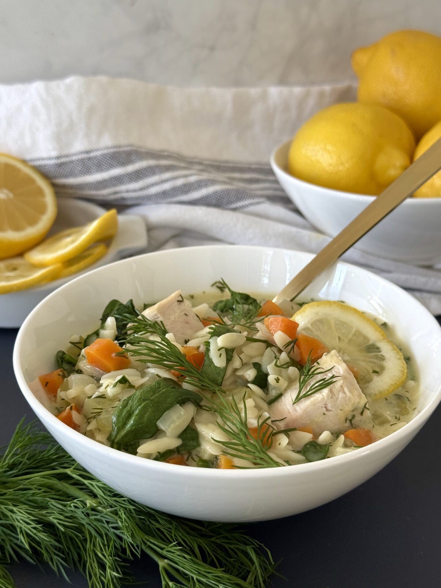 A bowl of Avgolemono soup is seen in the foreground, garnished with fresh dill. Bowls of lemons are seen in the background, along with a blue and white cloth napkin.