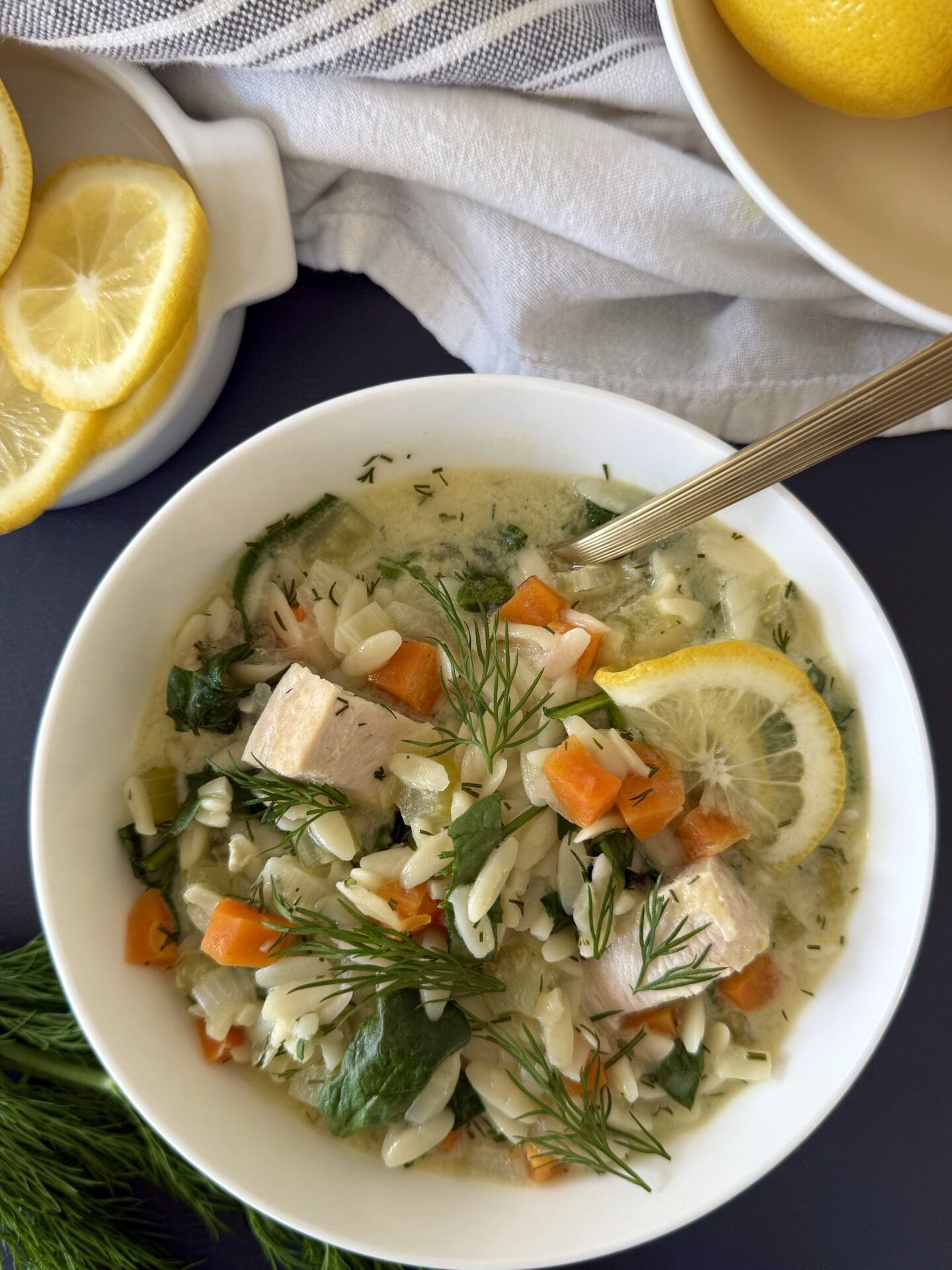 Avgolemono soup is seen from above, garnished lavishly with fresh fill and surrounded by bowls of lemons.