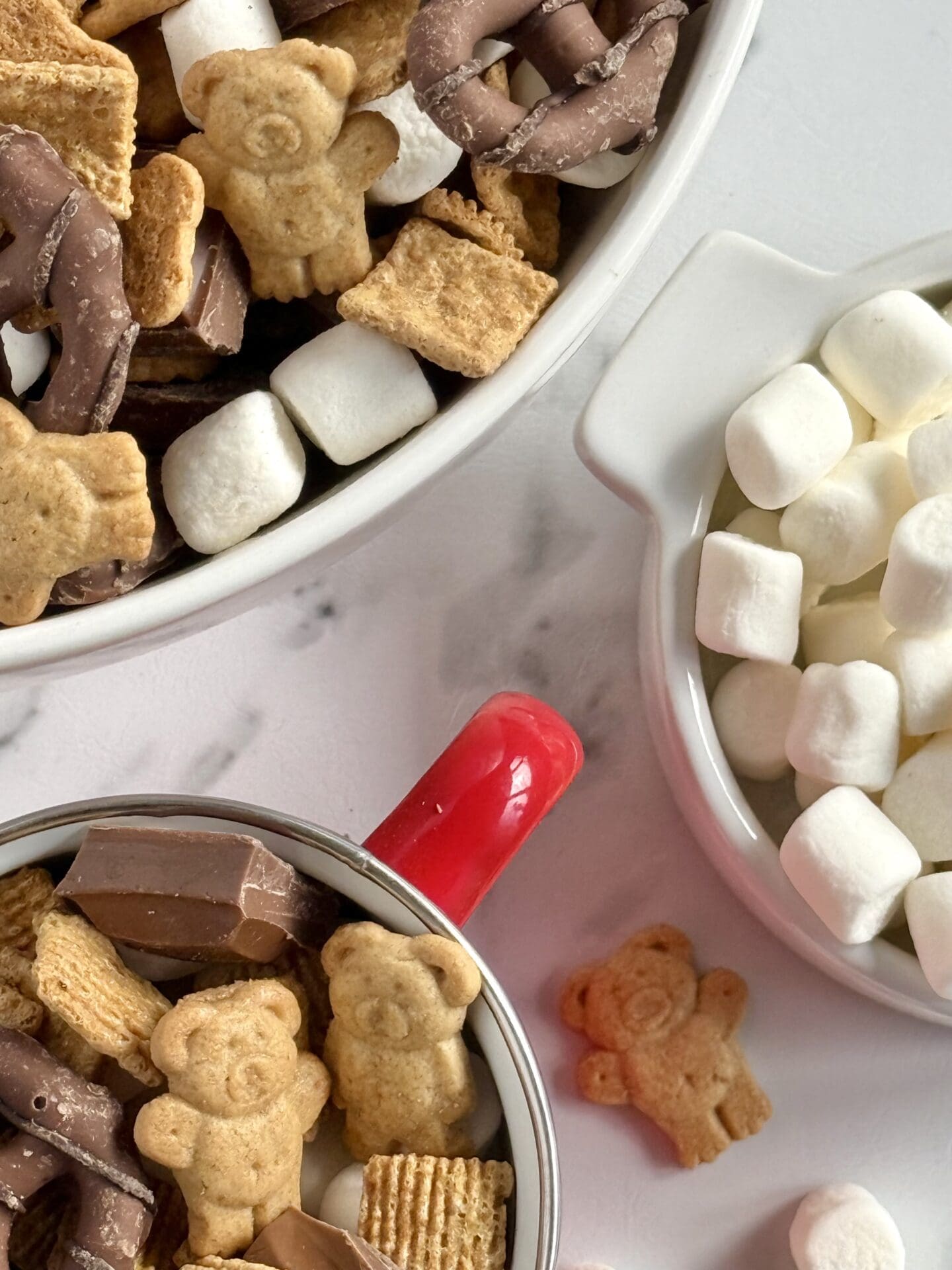 Three bowls of S’mores Snack Mix and mini marshmallows are seen from above.  Marshmallows and Teddy Grahams spill from the bowls onto the white marble counter. 