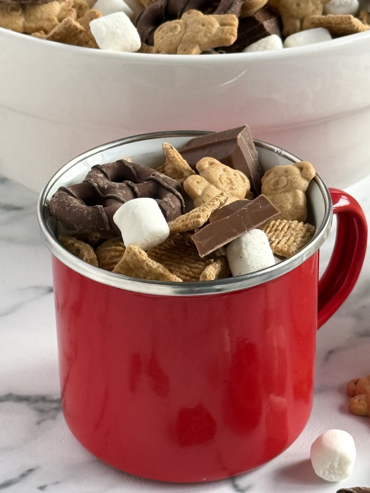 A red enamel mug is filled with S’Mores Snack Mix, for an easy, portable versions of classic campfire s’mores. 