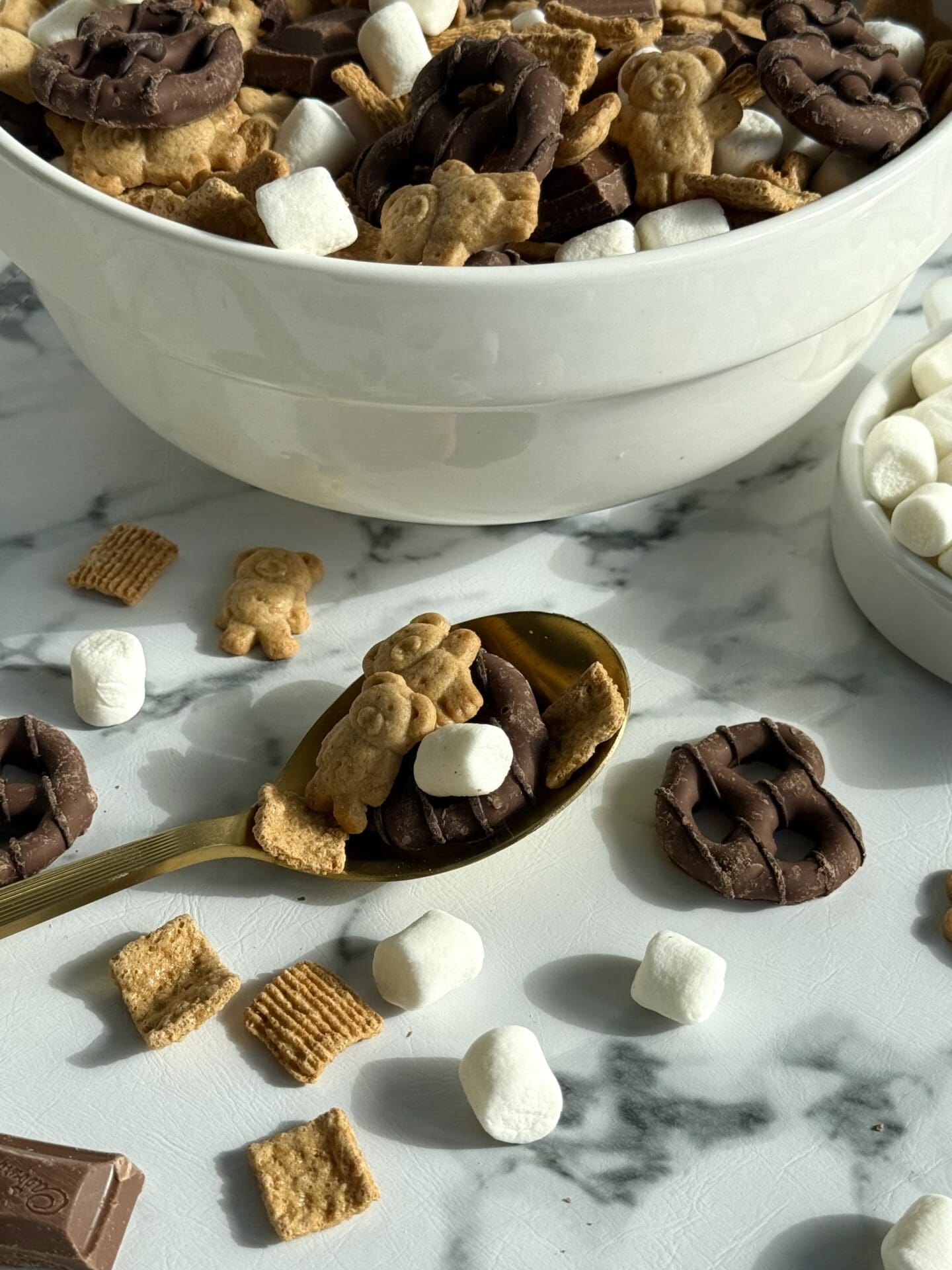 A large bowl of S’Mores Snack Mix sits on in the sunlight. A gold spoon appears in the foreground, spilling chocolate covered pretzels, marshmallows, Golden Grahams and chunks of milk chocolate onto the counter.