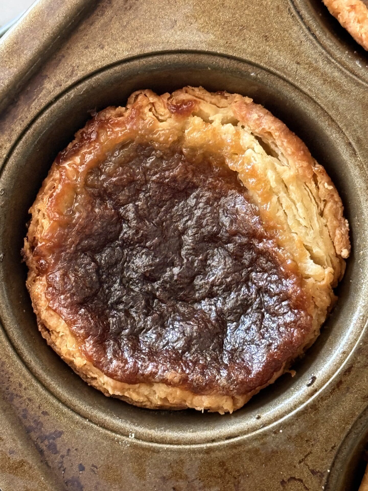 A single butter tarts is seen from above, sitting in its pan.  The flaky, golden layers of the pastry crust can clearly be seen along with the glossy, caramel-like filling.  