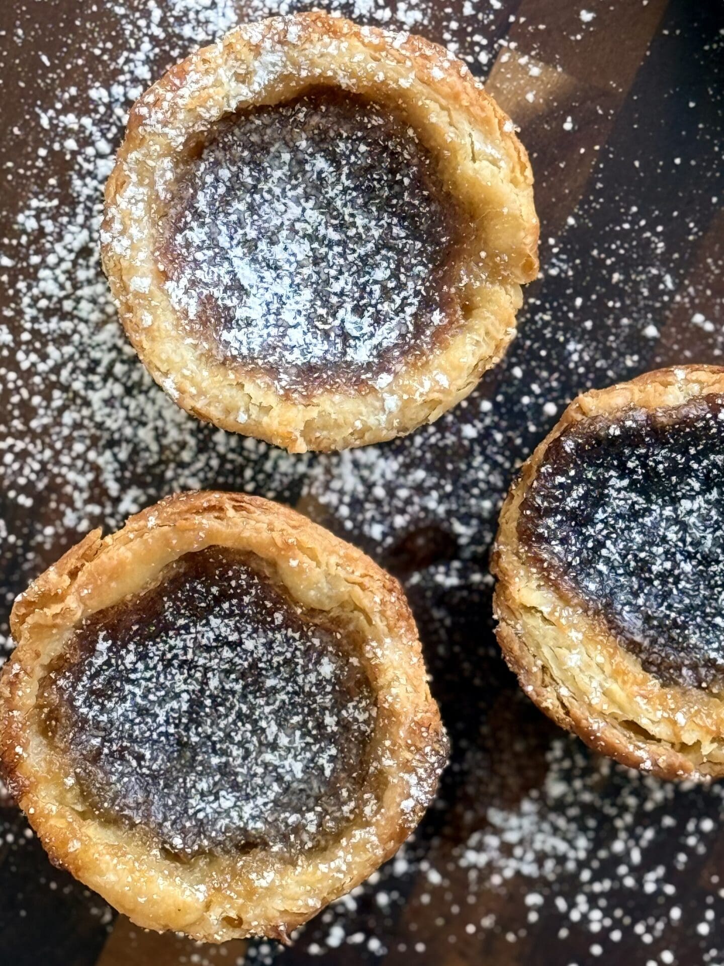 A trio of freshly baked butter tarts are seen from above, lightly dusted with powdered sugar.  The butter tarts show off the flaky, golden layers of their crusts and their silky, caramel-like filling