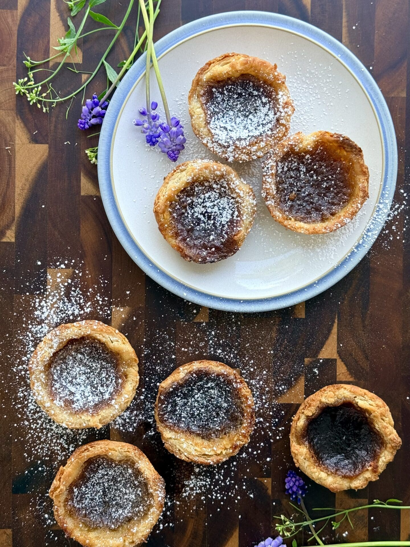 Freshly baked butter tarts are seen from above.  Some sit on a pretty blue and white plate, and some rest directly on a wooden board.  The tarts are dusted in confectioners sugar and surrounded by small purple and white flowers.