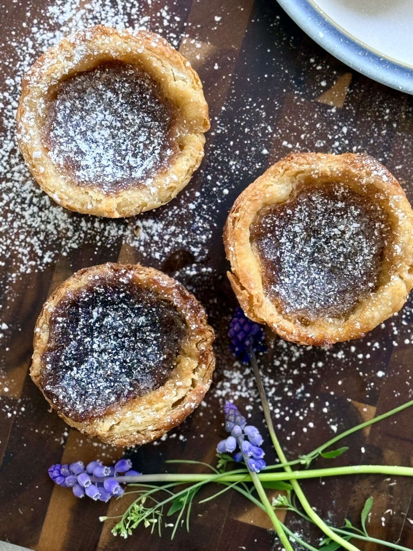 The Lakehouse Butter Tarts are seen from above, dusted in powdered sugar and resting on a wooden board.  Small purple and white flowers complete the image.
