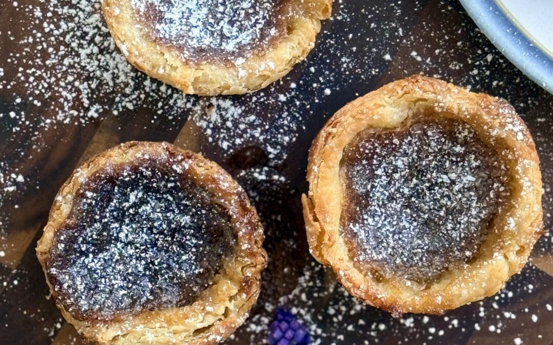 A collection of 3 freshly baked butter tarts sit on a wood board, dusted with powdered sugar and surrounded by small purple flowers.