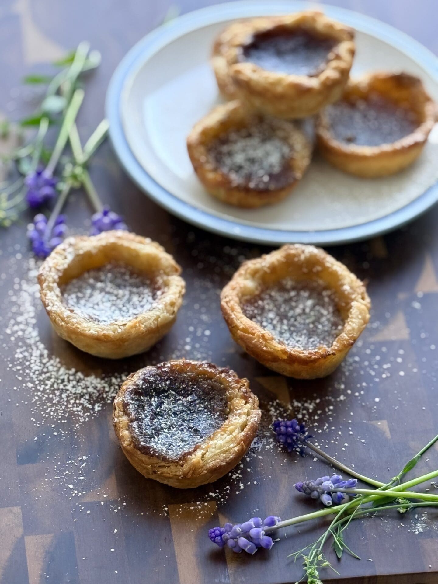 A trio of butter tarts sits in the foreground of the photo, on a wooden board.  In the background, a small blue and white plate holds another 3 tarts.  Small, purple flowers complete the scene.