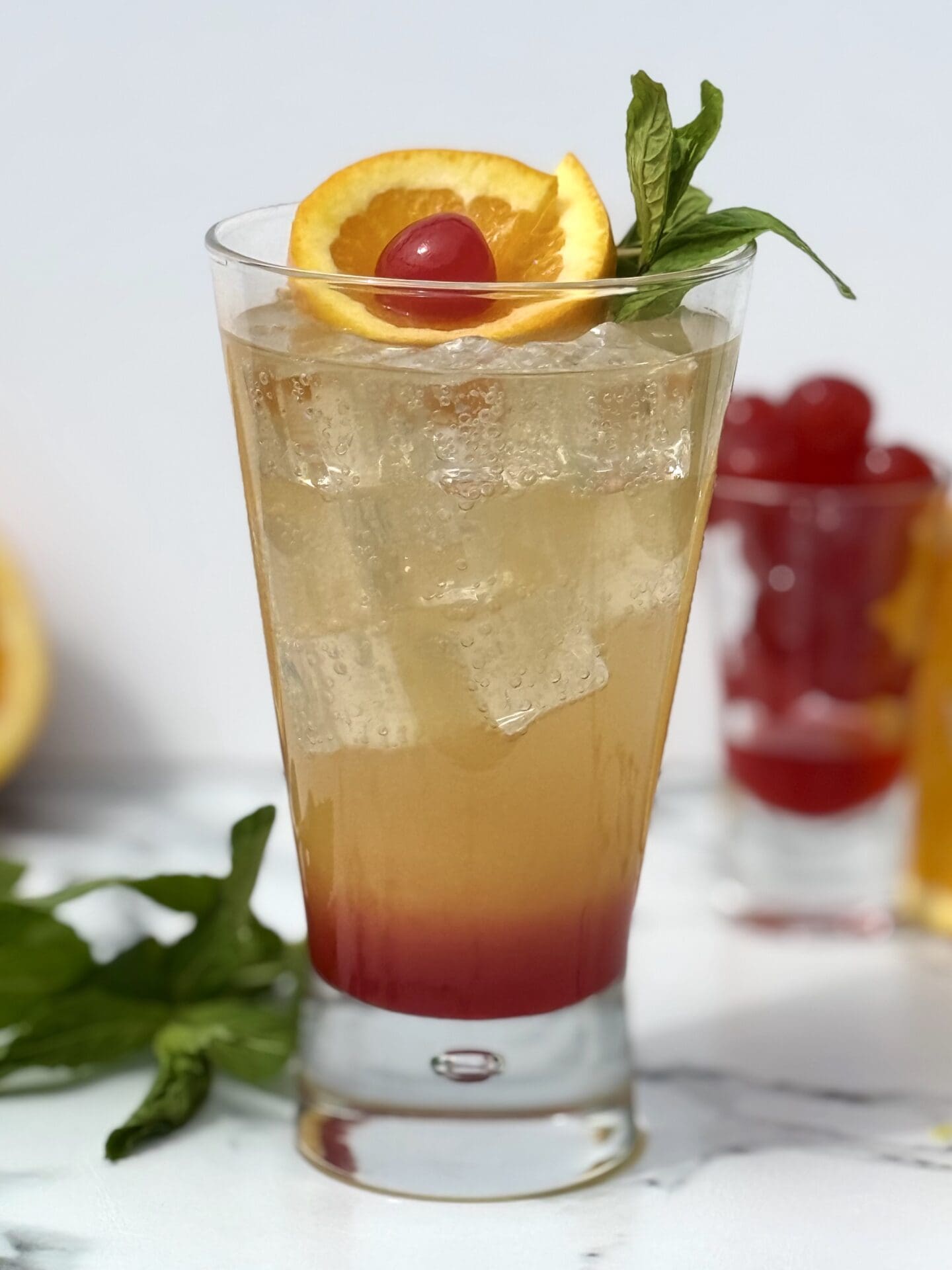 A Shirley Temple drink is seen in a tall glass, garnished with cherries, an orange slice and fresh mint.
