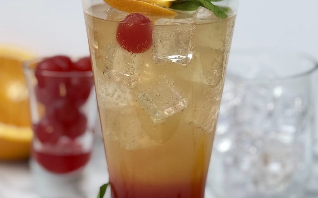 A Shirley Temple drink shows off its pretty layers of grenadine, orange juice and ginger ale, in a lavishly garnished glass.