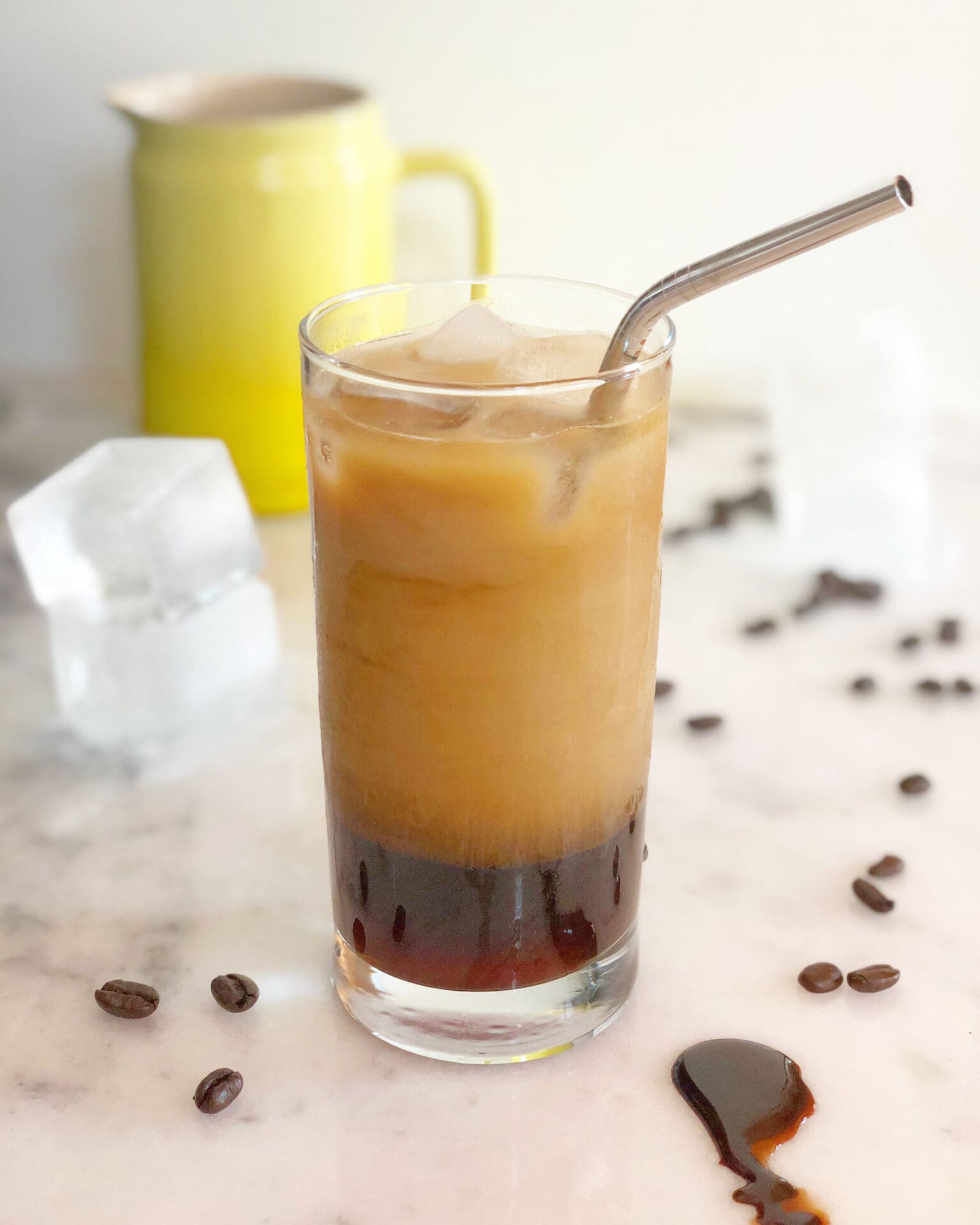 A glass of iced coffee sits in the foreground, showing off its layers of coffee and cream. The glass is surrounded by ice cubes, coffee beans and chocolate sauce.