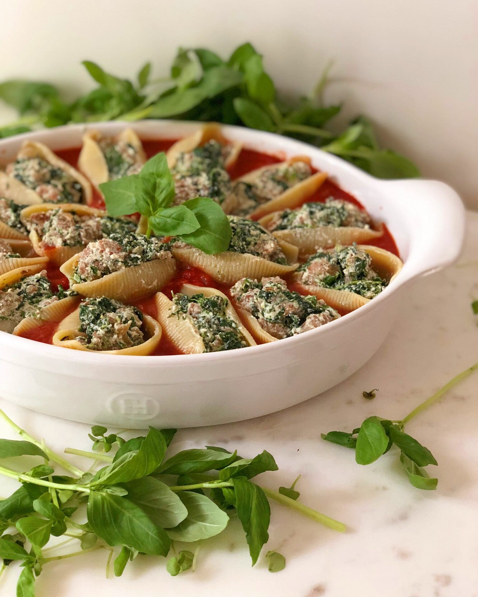 A white baking dish is filled with sausage, ricotta and spinach stuffed pasta shells, surrounded in marinara sauce and fresh basil.