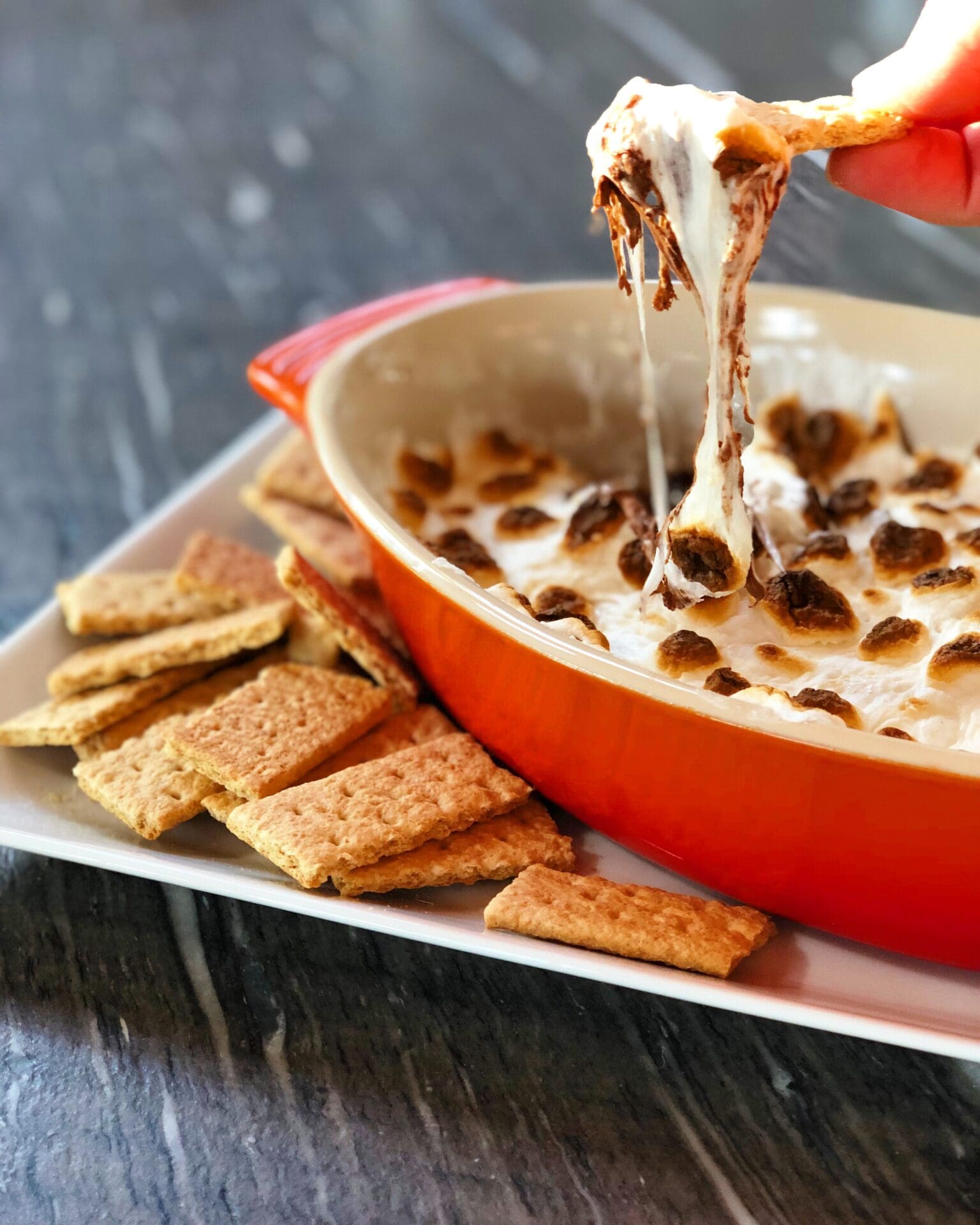 A dish of s’mores, baked in the oven, is surrounded by graham crackers.  A hand is seen dipping a cracker into the melted chocolate and marshmallows.  The marshmallows are gooey and stringy as the cracker lifts out of the dish.